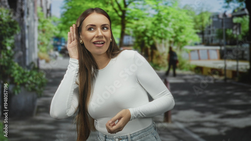 Young woman cupping her ear and pointing finger on a tree lined street near buildings, listening and gesturing for someone to speak; curiosity. Free Download Young woman cupping her ear and pointing finger on a tree lined street near buildings, listening and gesturing for someone to speak; curiosity. - GFXLoad