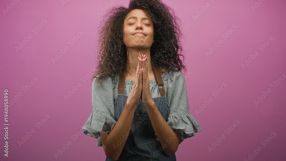 Obraz premium Woman with curly hair wearing denim apron, hands pressed together at chin in studio pink backdrop, closed eyes in praying gesture; hope.