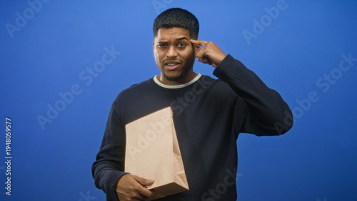 Fotografie Man holding a paper bag with finger to temple and furrowed brow in studio, facing an apparent delivery mixup and order delay; frustration