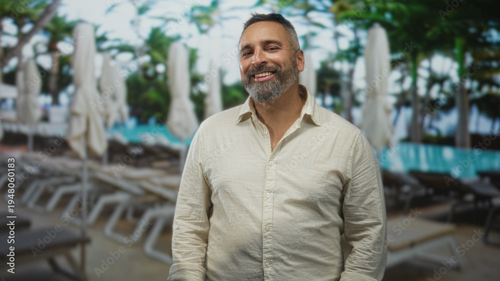 Naklejka premium Man standing with salt and pepper beard, hands relaxed at sides, neutral expression near pool loungers in building pool area; quiet contentment.