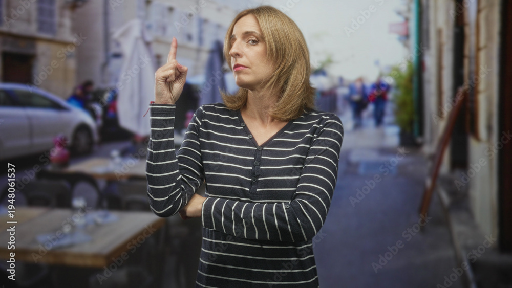 Fototapeta premium Woman points finger upward with folded arms at street cafe table, looking to the side; impatience urgency.