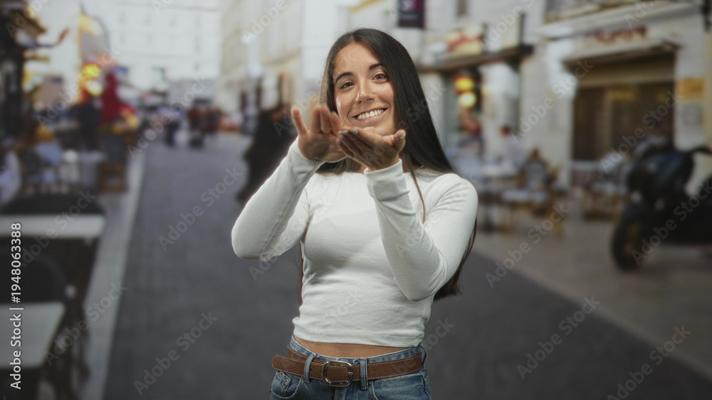 Fototapeta premium Young hispanic woman pinching fingers and smiling with visible hands and casual white top on a bustling pedestrian street lined with cafes and scooters; playful joy.