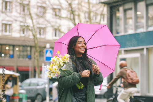 Happy young Latina woman holding flowers under pink umbrella on rainy city street, stylish female enjoying spring bouquet while walking in urban European setting
