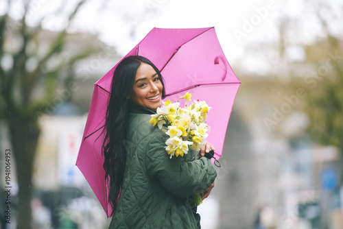 Smiling young Latina woman holding bouquet of yellow spring flowers under pink umbrella on rainy city street, happy female enjoying fresh daffodils during wet weather outdoors