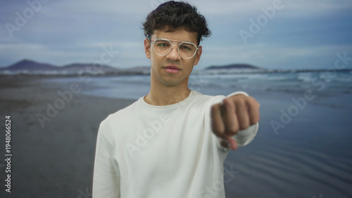 Man pointing finger at camera on beach standing against gentle waves and sandy shore wearing glasses and casual sweater; assertion.