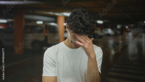 Young hispanic man pinches nose next to a parked car in an underground parking building; odor disgust.