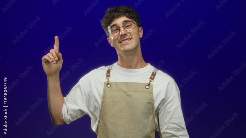 Naklejka premium Man pointing index finger in blue studio, smiling and wearing beige apron and round glasses; playful curiosity.