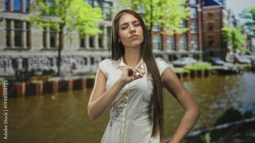 Obraz premium Young woman points finger near face beside canal building in city wearing white dress and long hair; contemplation.