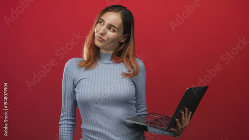 Photography Woman wearing blue turtleneck sweater, holding laptop with a head tilt in a red studio; thoughtful curiosity