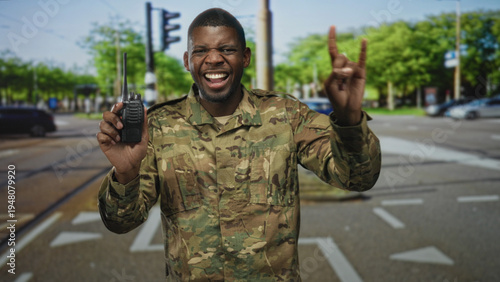 Konstfotografi Soldier man in camouflage uniform smiles, holds a walkie talkie and flashes rock horns while standing at a busy street intersection; pride duty