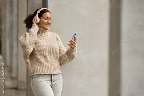 Young woman in a beige turtleneck sweater and headphones leaning against a concrete wall, smiling while using her smartphone. Modern urban lifestyle, cozy autumn fashion, and digital connectivity.