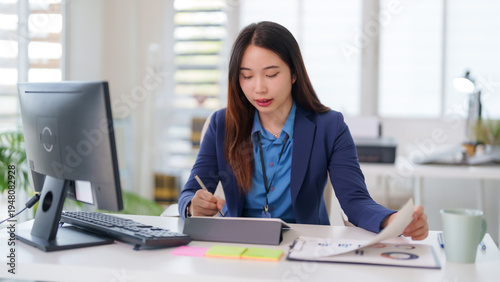 Asian adult business professional woman taking notes digital tablet analyzing business reports modern office desk workplace productivity