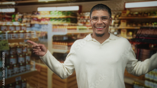 Man in cream sweater and glasses pointing with crossed arms at crowded store shelves of jars and bottles inside a grocery building; playful decision.