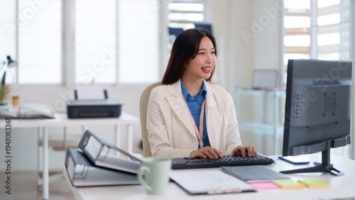 Asian adult business professional woman working computer typing keyboard managing tasks achieving goals bright modern office workstation productivity success