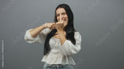Εκτύπωση καμβά Young hispanic woman in white blouse shows middle finger to camera while making a playful punching motion in a gray studio setting; defiance playfulness