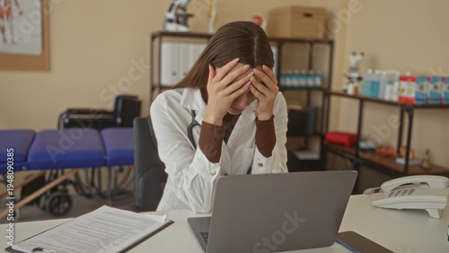 Foto Woman doctor in white coat with stethoscope holding hands to face at desk in building, laptop and clipboard on table; fatigue