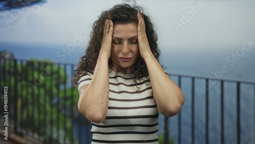 фотографія Woman presses temples with hands on street railing beside the sea under bright daylight; stress and fatigue