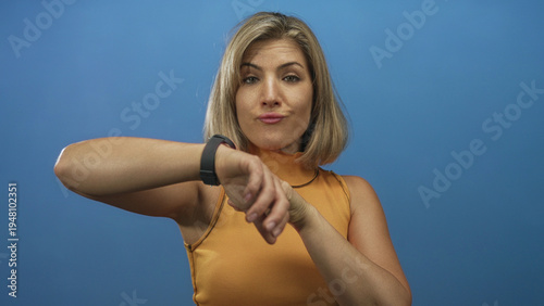 Quadro su tela Woman wearing orange sleeveless top pointing finger to watch on bare wrist in blue studio; impatience time pressure urgency