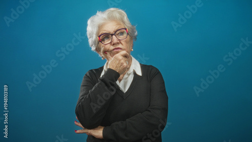Photos Elderly woman wearing glasses and a collared shirt with sweater, hand on chin and folded arm, looking upward against blue studio backdrop; pensive reflection