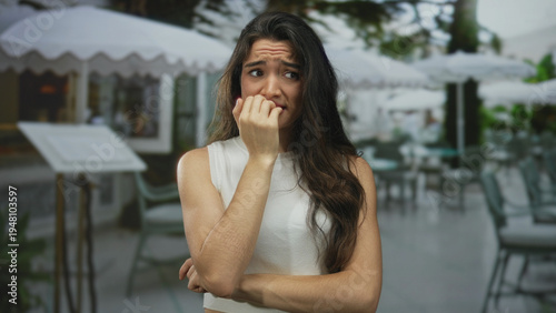 Photos Young hispanic woman biting fingernails with worried gaze on restaurant terrace; anxiety worry uncertainty