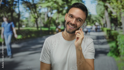 Young hispanic man smiling while talking on a cellphone in a vibrant green park with a blurred background and tree-lined walkway during a sunny outdoor day.
