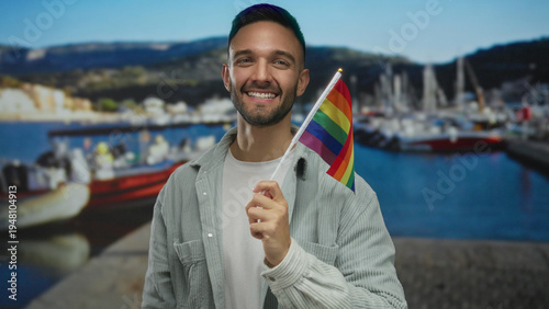 Canvas Print Young man smiling and holding rainbow flag on seaside promenade with boats in background under clear blue sky