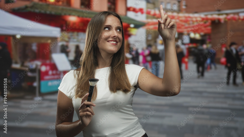 Fototapeta premium Woman holding a makeup brush and pointing index finger upward amid a busy street market with red lanterns and stalls; playful confidence.