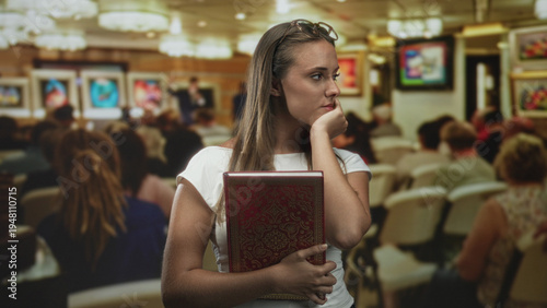 Fotografie Young brunette woman holding a red ornate book, hand to mouth gesture while standing among seated audience in auction building; pensive uncertainty reflection