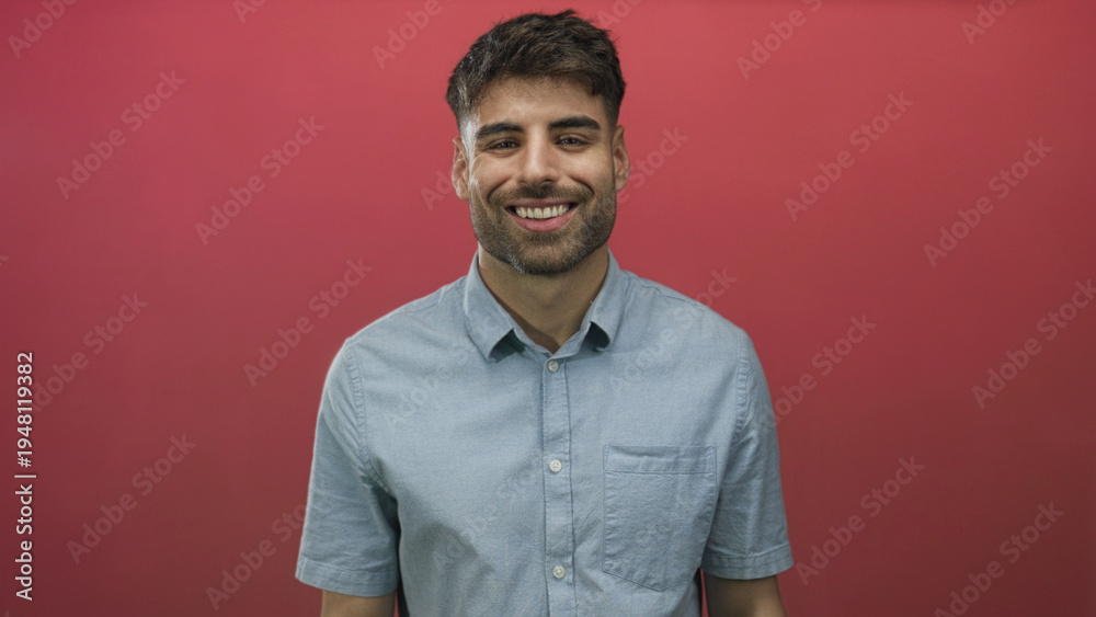 Obraz premium Young hispanic man smiling with teeth in red studio wearing light blue shirt; confidence joy optimism cheerful.