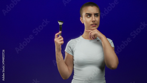 Konstfotografi Woman holding makeup brush with finger on chin in studio, short buzzed hair, white tee, examining brush near face against deep blue backdrop; uncertainty