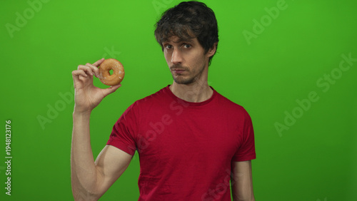 Cuadro en lienzo Man holds glazed donut up to camera at eye level in bright green studio with neutral expression; hunger craving desire appetite