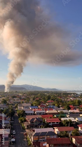 Large smoke plume rising over residential town as fire burns near neighborhood with mountains in background, thick pollution cloud spreading across sky above houses and streets in populated urban area
