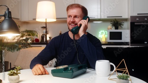 Young man at night home answers vintage wired telephone says hello smiles with nostalgia calm mood. Caucasian guy on sofa at table talks on corded phone drinks coffee laptop quiet calm evening gentle.
