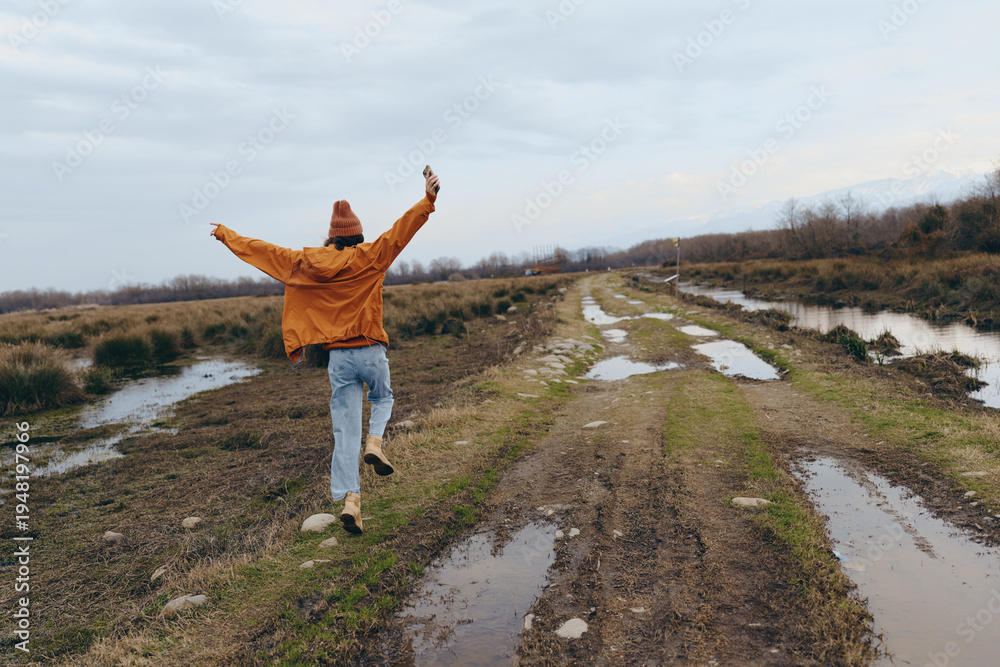 Fototapeta premium A woman lifestyle concept showing a joyful person in casual clothes walking on a muddy rural road with arms raised. The cloudy sky and natural landscape create a carefree outdoor mood.