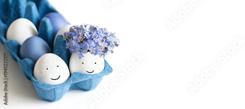 Easter eggs with painted faces and flowers in a blue cardboard box on white background. Copy space. Banner.