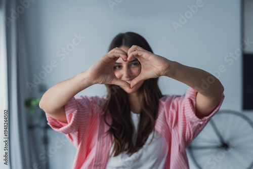 Attractive female showing a heart symbol with folded fingers, looking cheerful and confident, symbolizing friendliness, connection, and engagement in a casual indoor setting