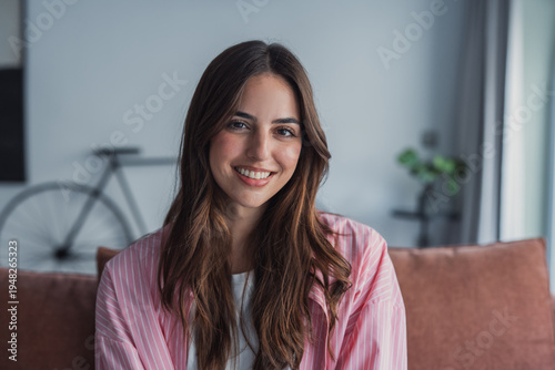Happy attractive young adult woman housewife sitting on sofa looking at camera in modern cozy home, smiling single 20s lady beautiful face relaxing on couch alone enjoy wellbeing posing for portrait