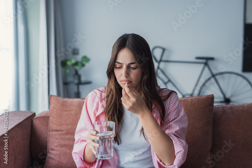 Quadro em tela Young woman at home holding a pill and glass of water, looking concerned while c