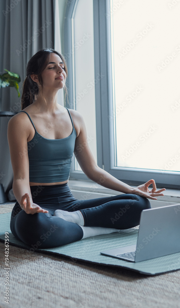 Fototapeta premium Young woman practicing yoga and deep breathing on a mat at home, following an online tutorial on her laptop, calm and focused, wellness, mindfulness, fitness lifestyle indoors