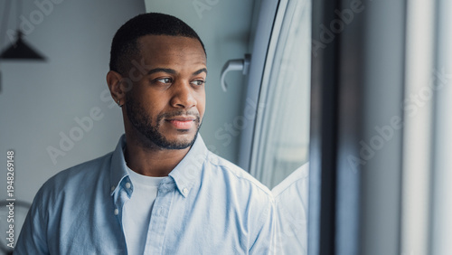 Fotografie Thoughtful millennial African American man looking at window away in deep thoughts, thinking over problems, bad news, difficult tasks, feeling bored, sad