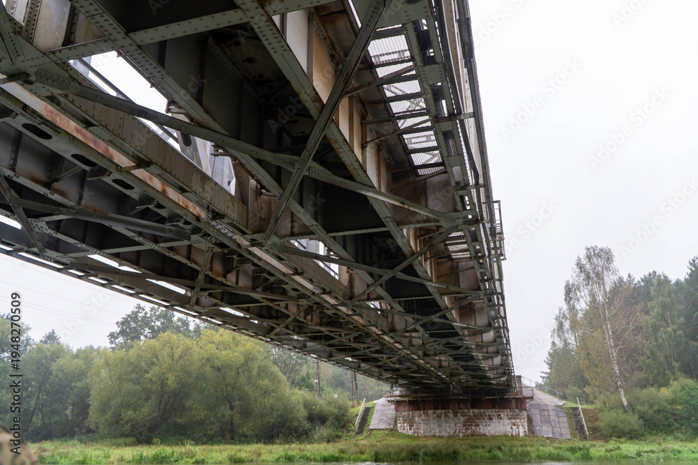 Fototapeta premium Wide angle perspective from below showing complex industrial steel girder network of railway bridge spanning river toward stone supports and forested shoreline under overcast sky.