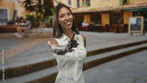 Fotografia Thai woman offers right hand for handshake on a street by a cafe building, smiling and leaning forward; friendly welcome