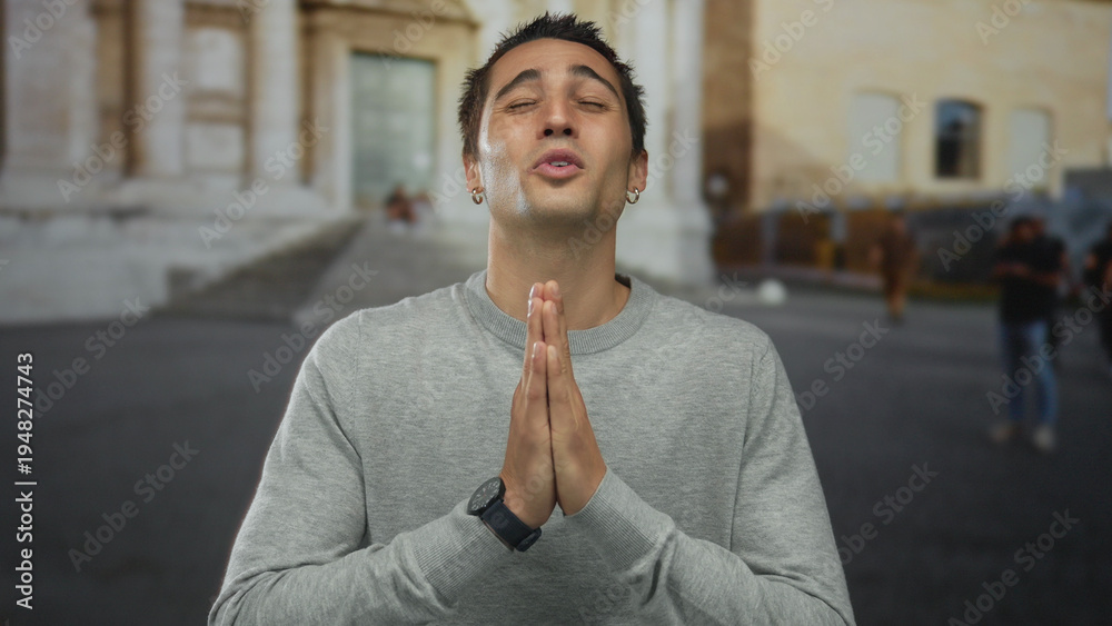 Naklejka premium Young man in old town outdoors, gesturing with hands in prayer near historical architecture, conveying hope and peace.