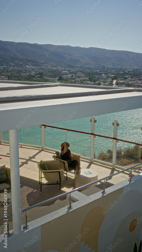 Naklejka premium Woman sitting holding sunhat by glass railing overlooking turquoise sea from cruise terrace building with wicker chairs and small table in sunlight; serenity.