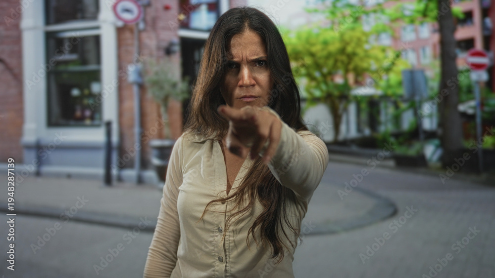 Fototapeta premium Woman points finger toward camera on street with furrowed brow and hand extended visible; anger confrontation.