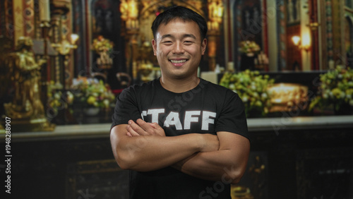 Fotografija Young chinese man smiling and wearing staff tshirt crosses arms in church building; service faith devotion