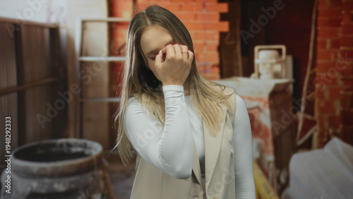 Canvastavla Woman indoors at construction site appearing worried with blonde hair wearing white top and beige vest surrounded by industrial equipment and unfinished walls displaying concern