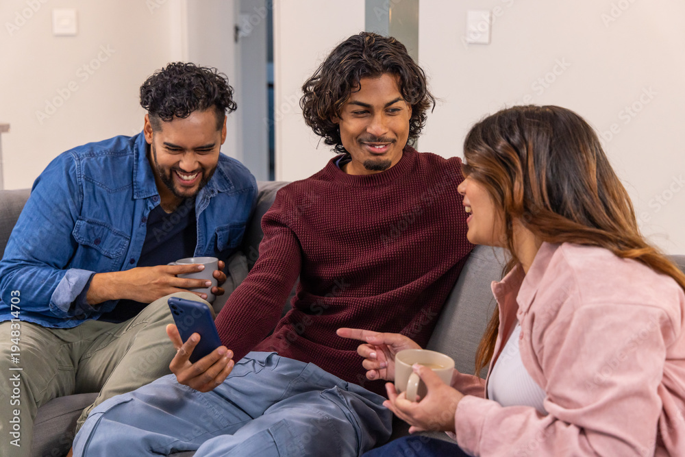 Fototapeta premium Asian friends in denim and sweaters laughing and pointing at smartphone on gray sofa with mugs