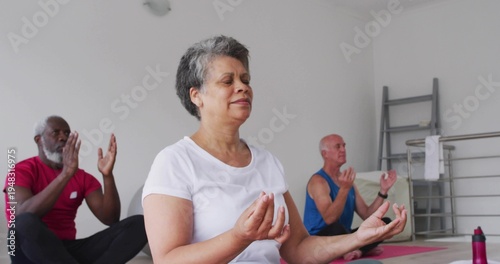 Sitting mature woman in white T-shirt breathing deeply in yoga studio, with mats and water bottle