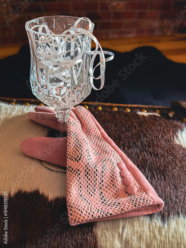 Wine glass with woman’s pink formal silver glove in soft leather sits on cow hide footstool. A bear rug & fireplace hearth are blurred in the distance.